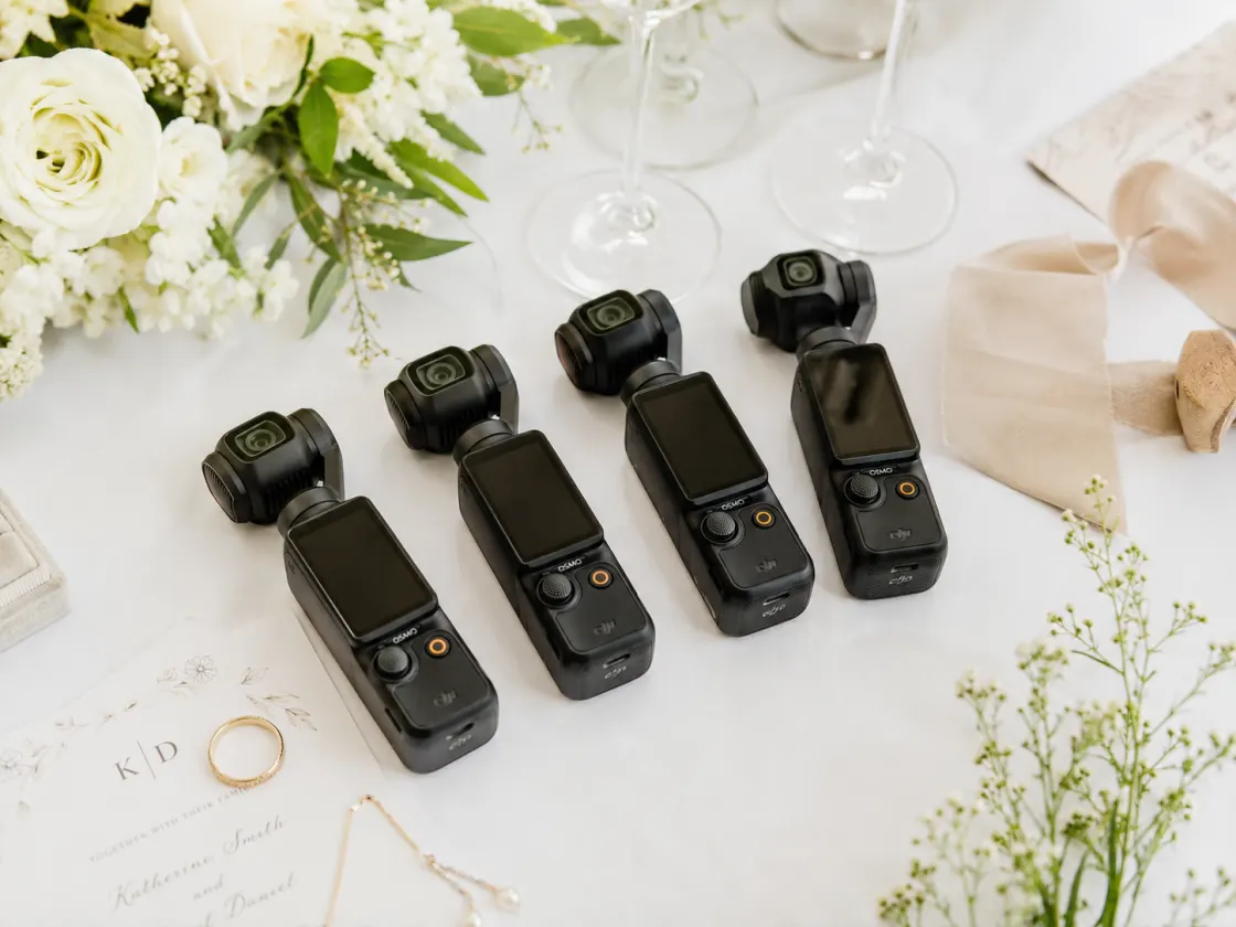 Four pocket cameras laid out on a white wedding table beside the flowers, the invitation and the rings