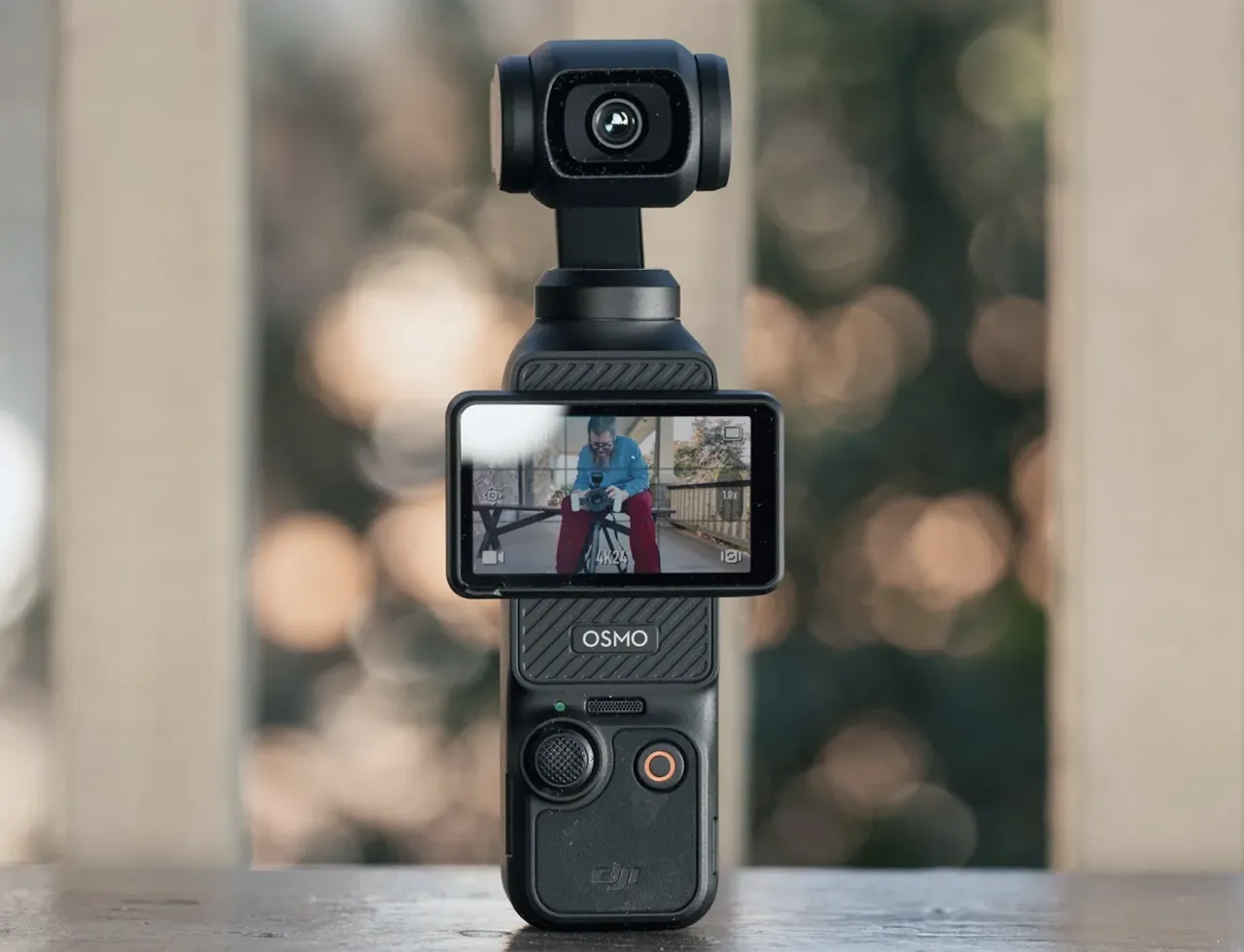 The camera standing on a table, screen on, with a blurred background behind it