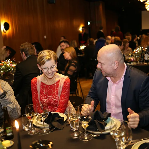 Two guests laughing at a dinner table as a room of others listens